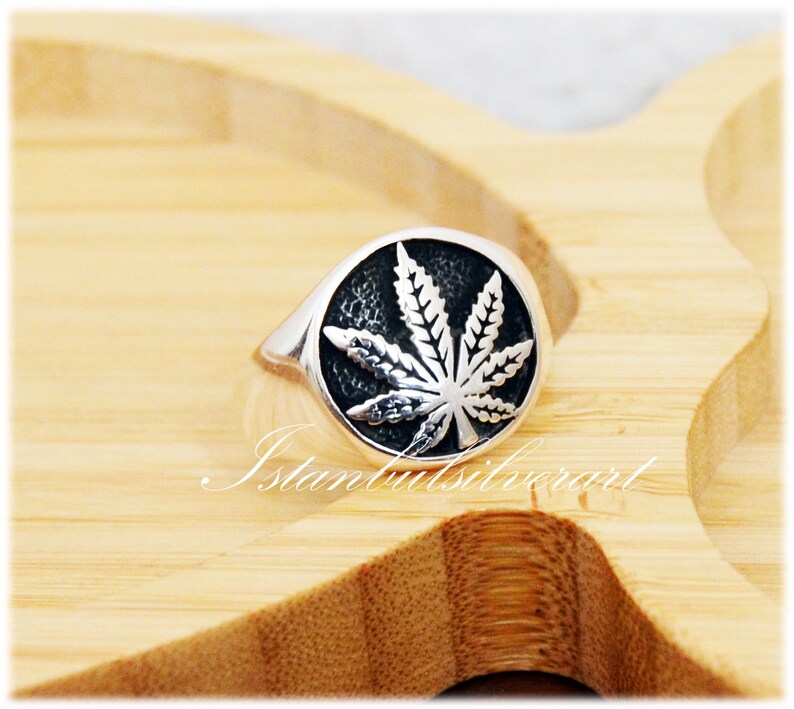 Puede incluir: Un anillo de plata con un dise&ntilde;o de hoja de marihuana negra. El anillo est&aacute; sobre una superficie de madera.