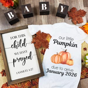 May include: A white baby onesie with a pumpkin graphic and the text "our little Pumpkin due to arrive January 2026." A framed sign reads "For this child we have prayed 1 Samuel 1:27." Wooden blocks spell "BABY."
