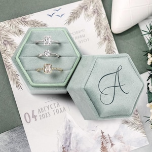 May include: A light green velvet hexagonal ring box with three rings inside. The rings have different stone shapes. The box is open, revealing the rings, and the lid is next to it. The lid has a cursive letter "A" on it.