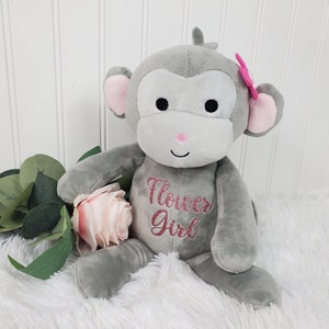 May include: A gray plush monkey with pink accents on the ears and nose, and a pink bow. The text "Flower Girl" is embroidered on the monkey's chest in pink glitter. A pink rose and green leaves are next to the monkey.