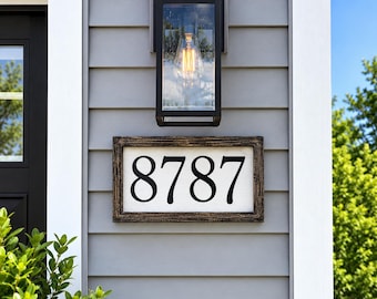 May include: A rectangular house number sign featuring the black digits 8787 on a white background, enclosed in a dark brown distressed wooden frame, mounted on gray horizontal siding beneath a black rectangular outdoor wall lantern light fixture.