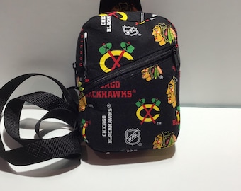 Może przedstawiać: Czarna torba na ramię z nadrukiem Chicago Blackhawks. Torba ma kieszeń zapinaną na zamek błyskawiczny i pasek. Torba jest wykonana z tkaniny i ma czarne tło z czerwonymi, żółtymi i białymi akcentami. Torba ma logo NHL i napis "Chicago Blackhawks".