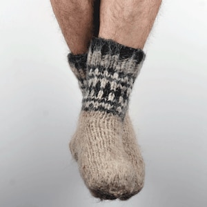 Può includere: Un paio di calzini di lana lavorati a mano. I calzini sono di colore beige chiaro con un motivo geometrico bianco e nero intorno alla caviglia. I calzini sembrano spessi e caldi.