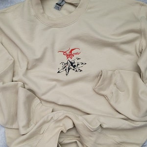 May include: A beige sweatshirt with a black and red embroidered design of a mountain with a dragon flying above it.