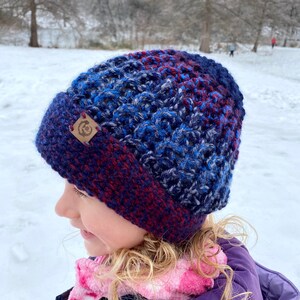 Puede incluir: Un niño con un gorro de punto azul, rojo y blanco con un parche de cuero marrón que dice "C". El niño lleva una bufanda rosa y blanca y una chaqueta morada. El niño está de pie en un campo nevado.