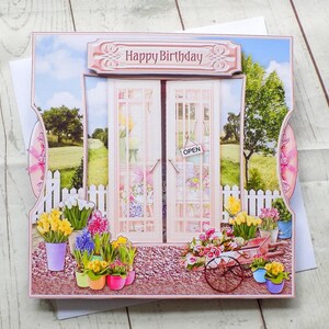 May include: A 3D birthday card featuring a garden scene with a sign that says "Happy Birthday." The card has a pair of doors with an "Open" sign, a white picket fence, and colorful potted flowers.
