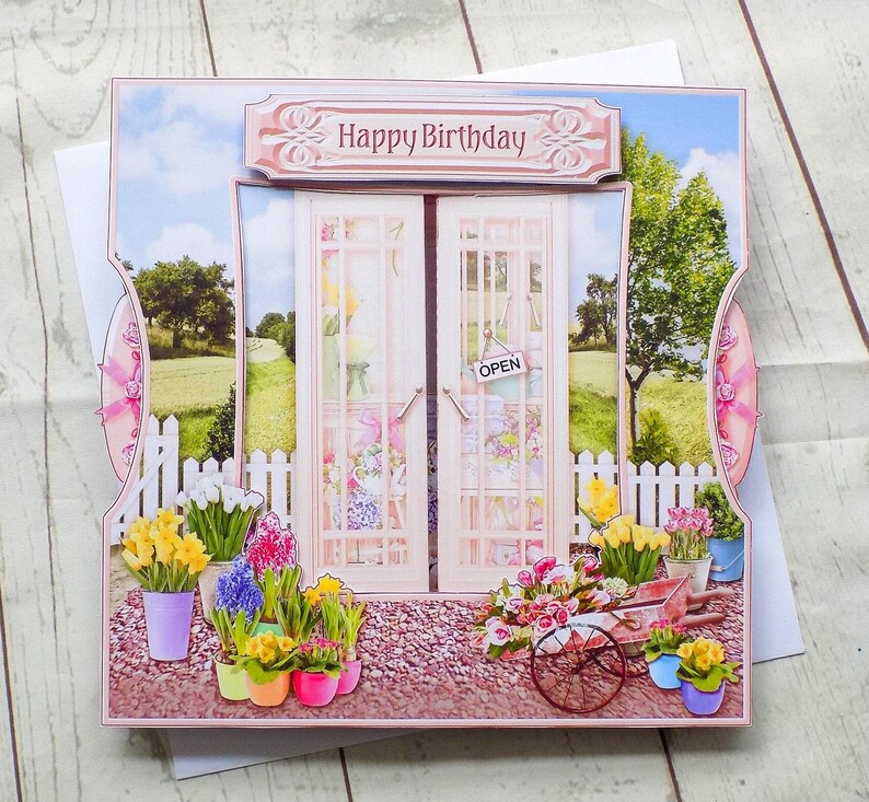 May include: A 3D birthday card featuring a garden scene with a sign that says "Happy Birthday." The card has a pair of doors with an "Open" sign, a white picket fence, and colorful potted flowers.