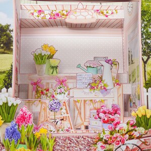 May include: A papercraft diorama of a flower shop, featuring a variety of colorful paper flowers, including tulips, daffodils, and hyacinths. The scene includes a watering can, a white picket fence, and a decorative cart, all in pastel colors.
