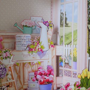 May include: A papercraft scene of a floral shop with a window overlooking a green landscape. The shop features various flower arrangements, watering cans, and decorative boxes. The color palette includes pink, green, and white.
