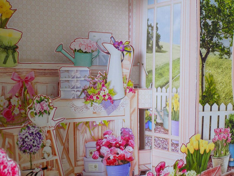 May include: A papercraft scene of a floral shop with a window overlooking a green landscape. The shop features various flower arrangements, watering cans, and decorative boxes. The color palette includes pink, green, and white.