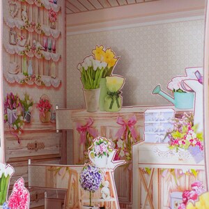May include: A pop-up paper craft scene of a floral shop with shelves, flowers, and a watering can. The scene includes white picket fences and a view of a green field and trees in the background. The shop is decorated with pink and white accents.