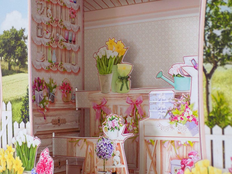 May include: A pop-up paper craft scene of a floral shop with shelves, flowers, and a watering can. The scene includes white picket fences and a view of a green field and trees in the background. The shop is decorated with pink and white accents.