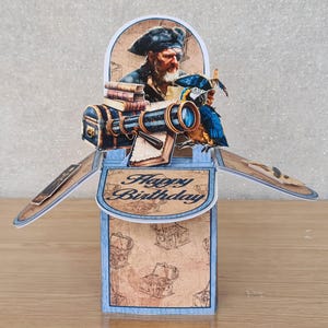 May include: A blue and brown pop-up card featuring a pirate holding a telescope and a parrot. The card is open and says "Happy Birthday" on the inside.