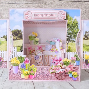 May include: A pop-up birthday card featuring a flower shop scene. The card has a pink and white color scheme with a sign that reads "Happy Birthday". The card is decorated with various potted flowers, a wheelbarrow, and a white picket fence.