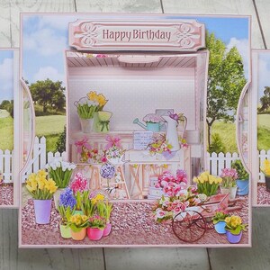 May include: A pop-up birthday card featuring a floral shop scene with the words "Happy Birthday" on top. The card has a pink border with floral accents and a garden scene background. Various potted flowers and a wheelbarrow are visible.