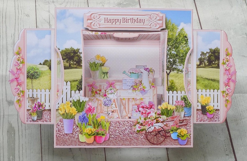 May include: A pop-up birthday card featuring a floral shop scene with the words "Happy Birthday" on top. The card has a pink border with floral accents and a garden scene background. Various potted flowers and a wheelbarrow are visible.
