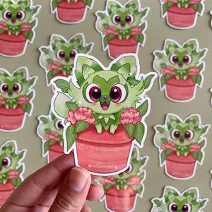 May include: A green and white cartoon cat with pink eyes and a pink nose sits in a red flower pot. The cat is surrounded by green leaves and pink flowers. The pot has a white heart with the text "aeriimart" on it.