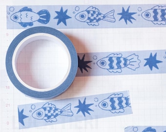 May include: A roll of blue washi tape with a repeating pattern of blue fish and stars. The tape is on a white gridded background.