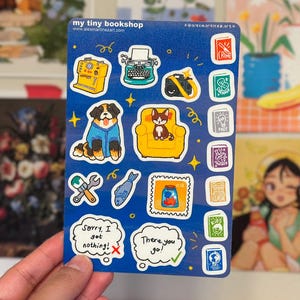 May include: A blue sticker sheet with various illustrations, including a sewing machine, typewriter, camera, dog in a blue sweater, cat on a yellow chair, and speech bubbles with text. The text on the sticker sheet reads "my tiny bookshop".