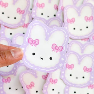 May include: A stack of purple and white bunny stickers with pink bows. The stickers are in the shape of a bunny with a fluffy tail and ears.