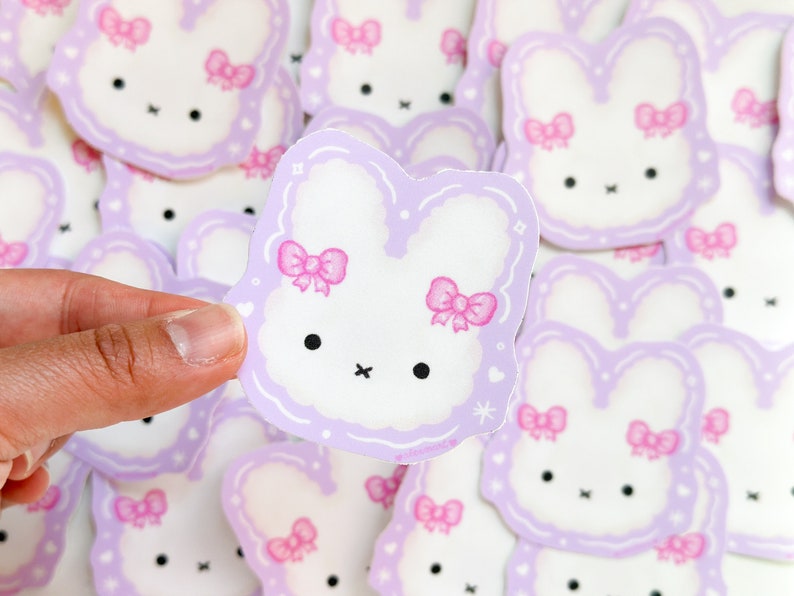 May include: A stack of purple and white bunny stickers with pink bows. The stickers are in the shape of a bunny with a fluffy tail and ears.