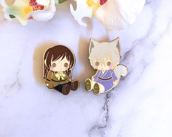 May include: Two enamel pins featuring anime characters. One pin depicts a girl in a brown uniform with a yellow bow. The other pin is a white fox-like character in a purple kimono. Both pins have gold-coloured metal borders.