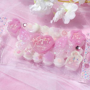 Op de afbeelding: Een roze en witte Nintendo Switch-hoes met een hartvormig ontwerp en de woorden "Soft Fairy". De hoes is versierd met glitter, strass en andere versieringen.