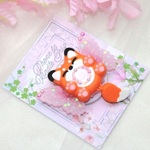 May include: An orange fox-shaped phone grip with black ears and white accents, featuring a white-tipped tail and pink glittery wings. The grip is on a pink card with the text "Phone & Kindle Grip".