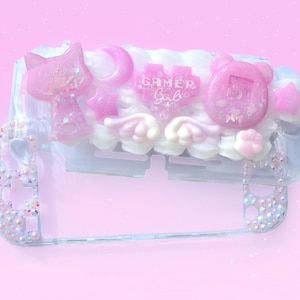 May include: A clear plastic Nintendo Switch case decorated with pink and white 3D embellishments. The embellishments include a cat, a star, clouds, wings, a heart with the text "Gamer Girl", and a pink clock with the number 15. The case is also decorated with iridescent rhinestones.