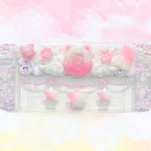 Op de afbeelding: Een doorzichtige Switch-consolehoes versierd met roze en witte versieringen. De hoes heeft een roze beer-, ster- en wolkenontwerp, met een roze en witte kralenketting. De randen van de hoes zijn ingelegd met iriserende strass-steentjes.