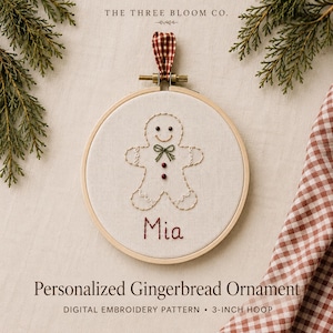 May include: A round wooden embroidery hoop containing a white fabric panel with a hand-stitched gingerbread person design. The figure features a green bow, two red buttons, and the name Mia embroidered in red thread below the figure.