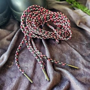 Puede incluir: Un par de cordones negros y rosas con aglets plateados. Los cordones están hechos de un material tejido y tienen un patrón a cuadros.