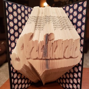 Könnte beinhalten: Ein Buch, das in die Form des Wortes "Arizona" gefaltet ist, mit einem Baseball-Motiv auf dem Einband. Der Einband ist blau mit weißen Baseballs.