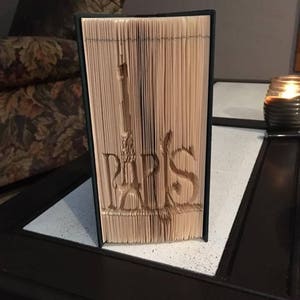 May include: A book with the word "PARIS" folded into the pages to create a 3D sculpture of the Eiffel Tower. The book is sitting on a wooden table.