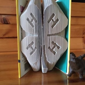 May include: A book folded into the shape of a four-leaf clover, with the letters "H" and "4" visible on each leaf. The book is standing upright on a wooden surface, with a green book behind it. The text "The Book Chiseler" is visible at the bottom of the image.