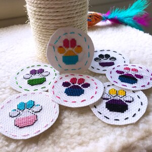 Puede incluir: Siete parches bordados con forma de huella de pata en varios colores, incluyendo las banderas del orgullo arcoíris, bisexual, transgénero y no binario. Los parches son circulares y tienen un fondo blanco con un borde cosido.