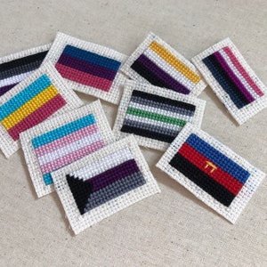 May include: A collection of 10 small, hand-stitched flags representing various pride flags. The flags are made with colorful embroidery thread on white fabric.