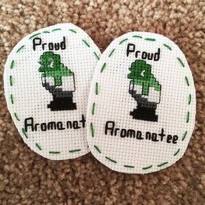 May include: Two white oval-shaped embroidered patches with green stitching around the edges. Each patch features a green pixelated figure with the text "Proud" and "Aromantic" in black.