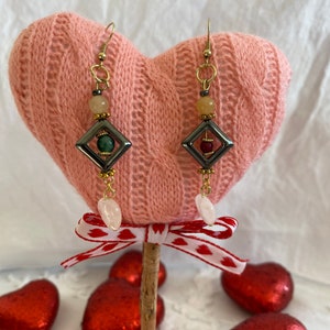 May include: A pair of gold earrings with a geometric design. The earrings feature a square frame with a green, red, and brown stone inside. The earrings are hanging from a gold hook and have a white stone at the bottom.