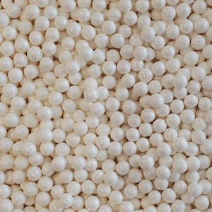 Puede incluir: Una imagen de primer plano de una gran cantidad de granas redondas blancas perladas. Las granas están dispuestas en un patrón aleatorio y llenan todo el marco.