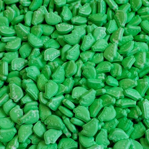 Puede incluir: Primer plano de un montón de caramelos verdes con forma de lima. Los caramelos son de un color verde vibrante y tienen un acabado ligeramente brillante. Los caramelos están esparcidos al azar, llenando el encuadre.