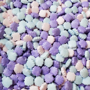 Puede incluir: Primer plano de un montón grande de espolvorear con forma de concha de mar de color pastel. Las chispas son en tonos de azul claro, lavanda y rosa.