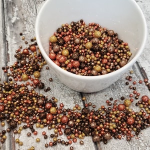 May include: A white bowl filled with a mixture of brown, gold, and copper coloured sprinkles. The sprinkles are scattered around the bowl on a wooden surface.