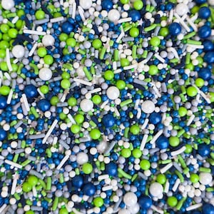 May include: A close-up of a colourful mix of sprinkles. The sprinkles include white, green, blue, and grey spheres, rods, and star shapes. The sprinkles are a mix of sizes and shapes, creating a textured appearance.