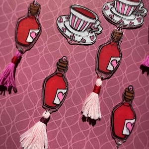 May include: Acrylic charms of a red potion bottle with a heart label and a pink tassel, and teacups with pink stripes and heart-shaped saucers. The charms are set against a pink patterned backdrop.