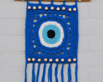 May include: A square crochet wall hanging featuring a central evil eye motif in black, light blue, and white, surrounded by a deep blue square pattern. The piece is suspended from a wooden dowel with light-colored wooden beads and blue fringe.