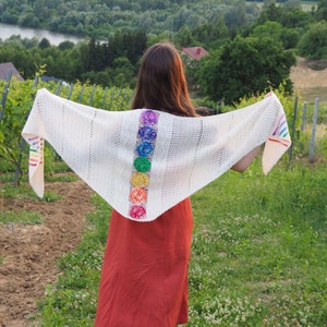 May include: A white crochet shawl with a rainbow design. The shawl is worn by a person standing in a field.