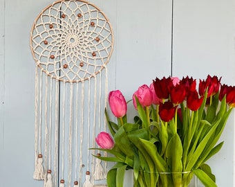 May include: A circular macrame wall hanging featuring a cream-colored woven web with brown wooden beads, suspended from a wooden hoop with long fringe tassels. Beside it sits a glass vase filled with red and pink tulips and two yarn spools.