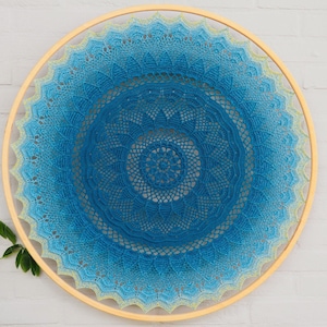 May include: A circular crochet wall hanging featuring a gradient of blue and teal yarn colours. The intricate, mandala-style pattern is stretched tightly within a smooth, light-coloured wooden embroidery hoop, creating a decorative fibre art piece.