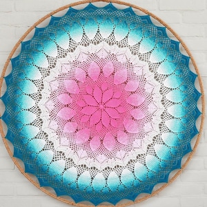 Op de afbeelding: Een rond, handgemaakt gehaakt stuk met een kleurovergang. Het midden is felroze, overgaand in wit, dan turquoise en uiteindelijk een diepblauwe rand. Het geheel is omlijst door een natuurlijk gekleurde houten hoepel.